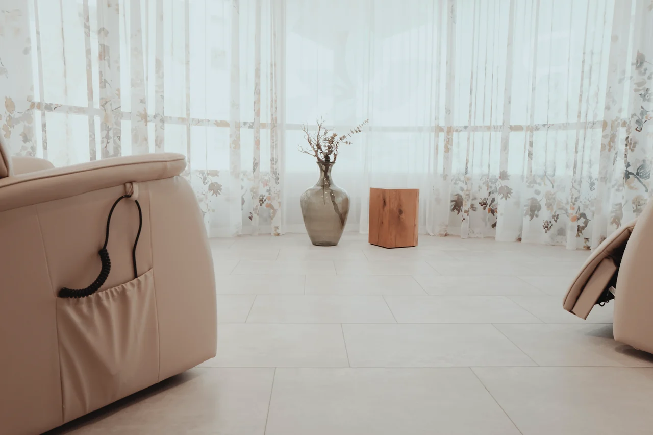 Leerer Raum mit cremefarbenen Liegesesseln und Vase vor Fenstern mit Blumenvorhang