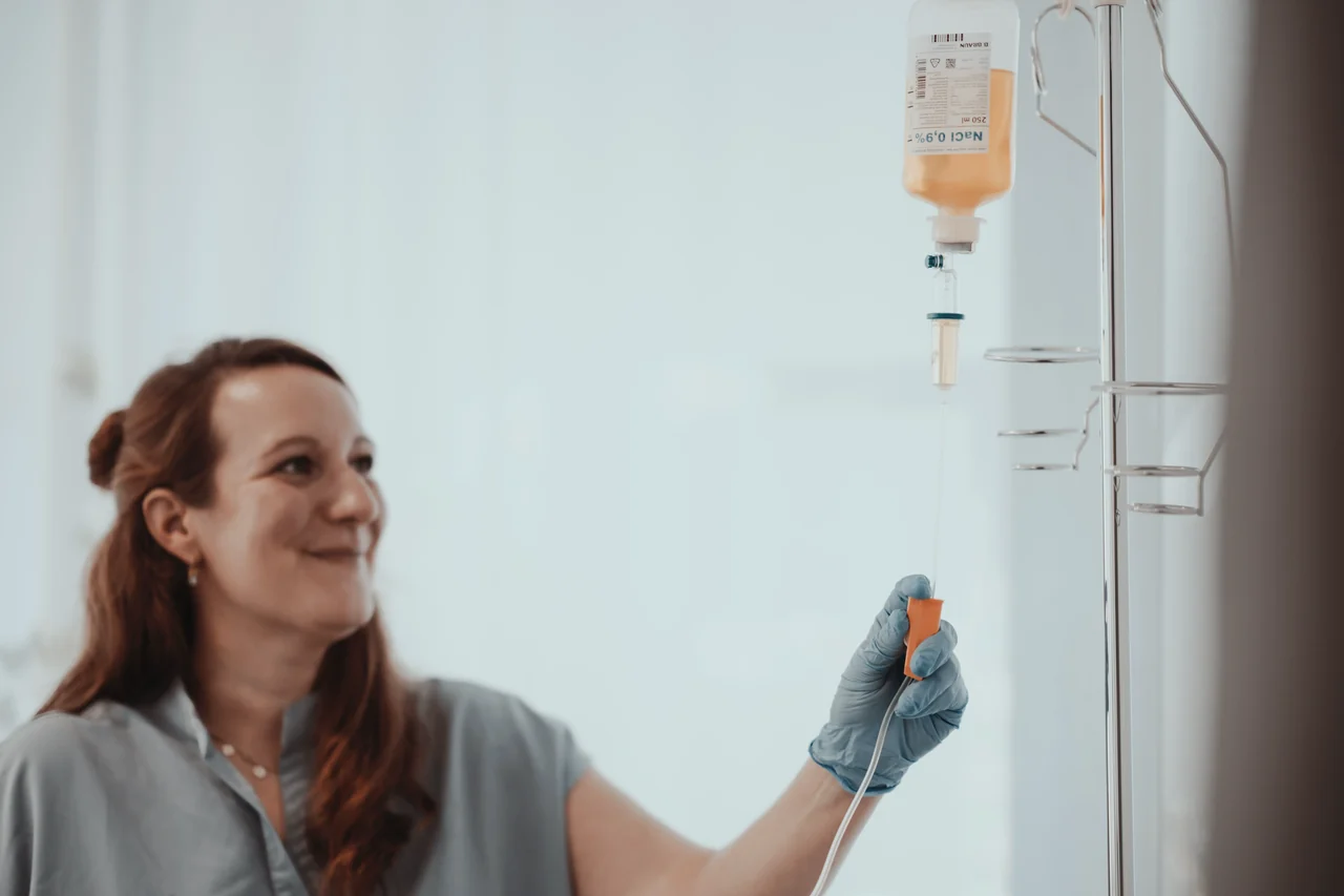 Mitarbeiterin hängt lächelnd einen Infusionsbeutel an einen Tropfständer.